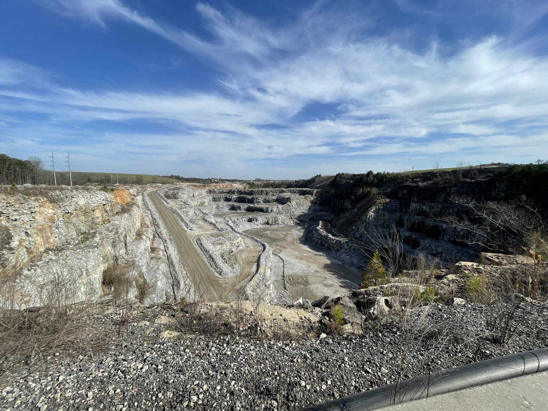 A wide view of a rocky quarry under a bright blue sky with scattered clouds. The landscape features layers of exposed stone and a dirt road winding through the site. Sparse vegetation is visible along the edges.