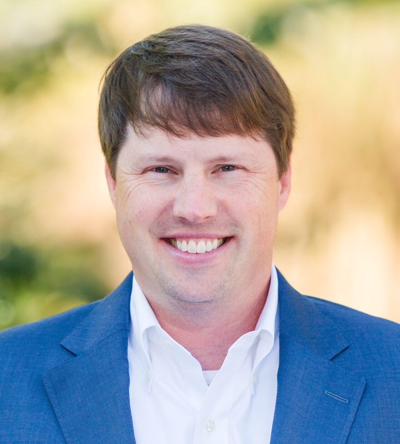 A man with short brown hair is smiling at the camera. He is wearing a blue suit jacket over a white shirt. The background is blurred with green and beige tones.