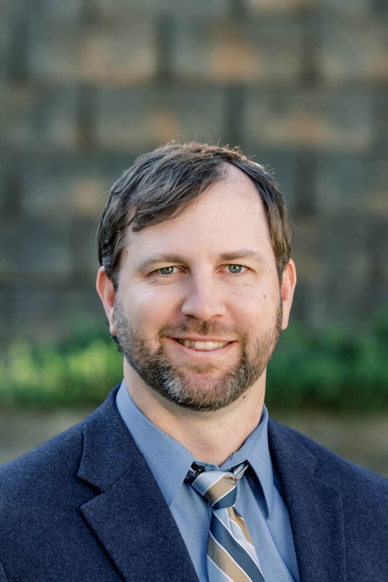 A man with short brown hair and a beard wears a blue suit jacket, blue shirt, and striped tie. He is smiling slightly and standing outdoors with a blurred stone wall and greenery in the background.