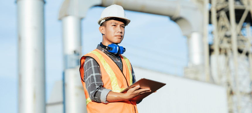 A construction worker in a hard hat, orange safety vest, and blue ear protection around his neck holds a tablet while standing at an industrial worksite.