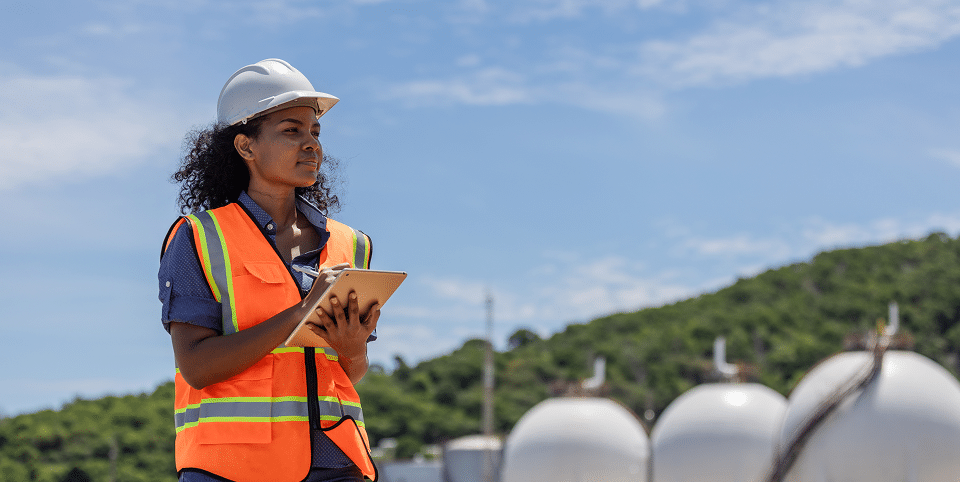 A worker wearing a hard hat and safety vest stands outside, holding a tablet and gazing forward. Behind them are industrial tanks set against green hills with a blue sky overhead.